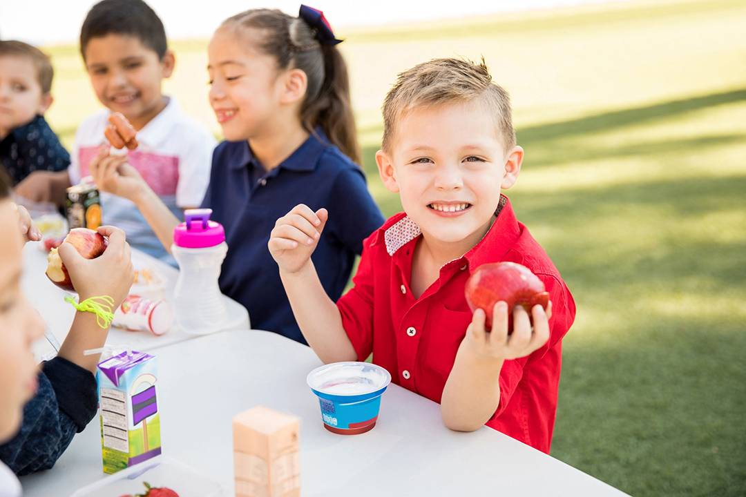 Adorable Caucasian preschooler enjoying his lunch with some of his friends at school and smiling