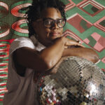 Artist Cauleen Smith posing with a disco ball in front of a geometric colorful backdrop