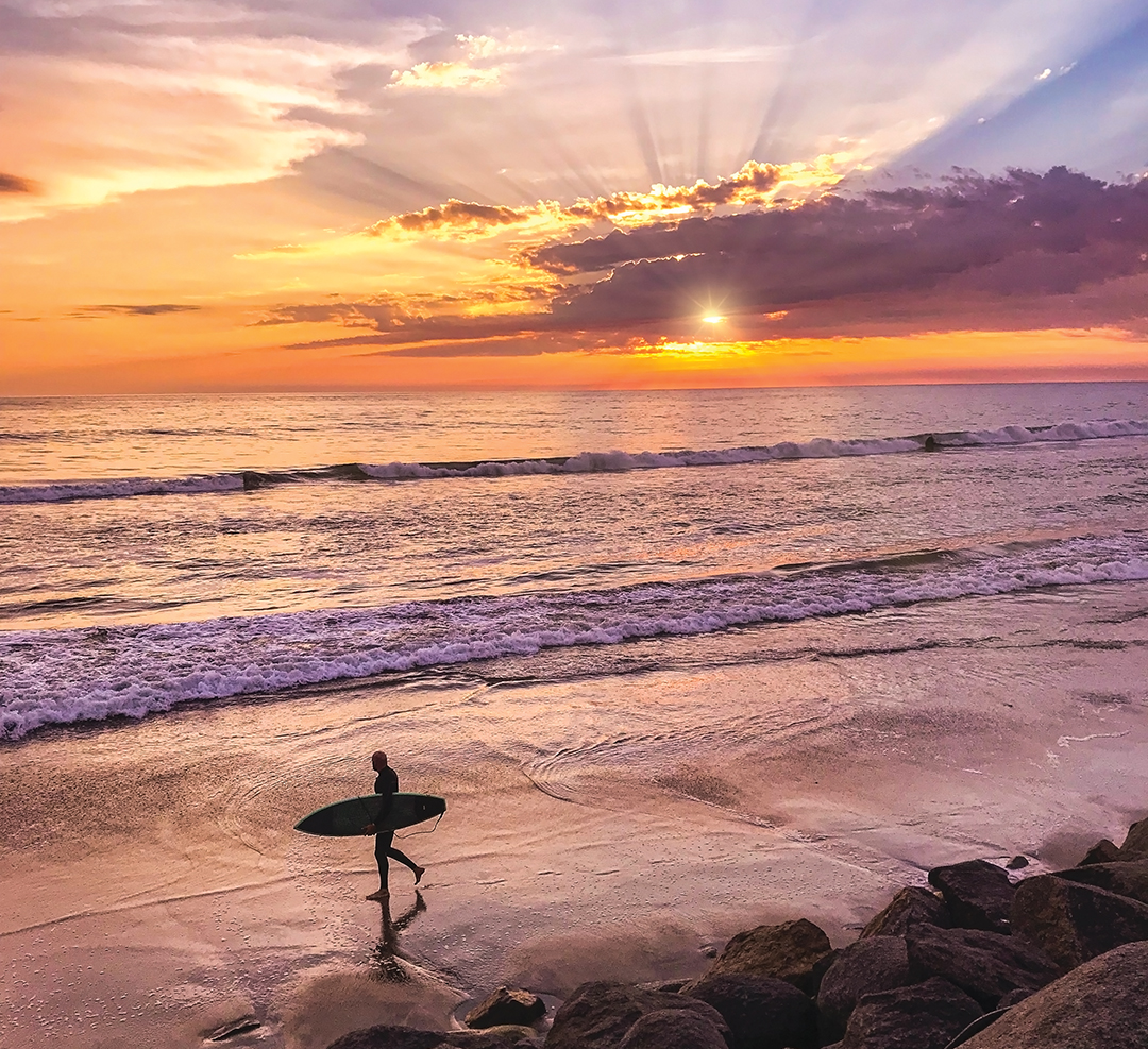 Silhouette of a surfer walking on the beach with a beautiful sunset on the water behind him
