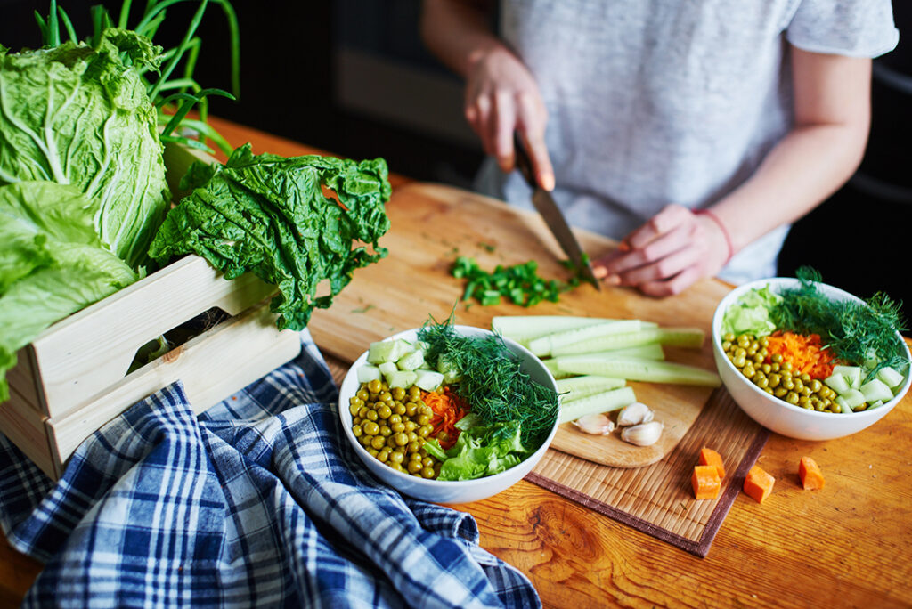Bowl with green peas, cucumbers, carrots, lettuce and dill standing on a table on a background of hand chopping green onions with knife on wooden Board