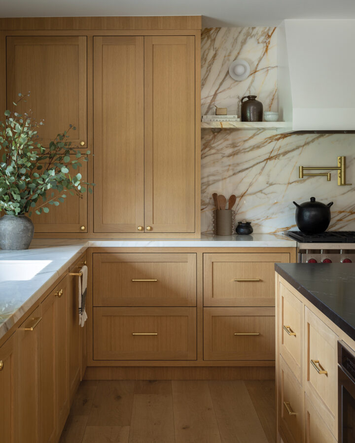 Valletta redesigned the kitchen for functionality, removing drop-down soffits, moving a desk, and rearranging the refrigerator and freezer. Warm white oak cabinets and a striking marble backsplash with black-and-gold veining above the cooktop became the kitchen’s focal point